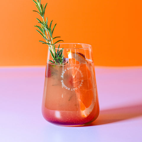 Glas der "Baby Got Drinks"-Kollektion, gefüllt mit einem bunten Drink mit Grapefruitstücken und Rosmarinzweig, auf lilafarbener Unterlage vor orangefarbenem Hintergrund.