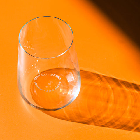 in einzelnes Glas der Kollektion „Baby Got Drinks“ auf orangefarbenem Untergrund. Die Glasgravur ist im Licht sichtbar und erzeugt einen eleganten Schattenwurf.