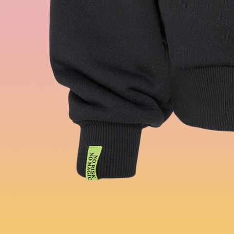 Ärmelabschluss eines schwarzen Sweaters mit geripptem Bündchen und neon-grünem Etikett mit schwarzem Text "NO RISK NO MAGIC".