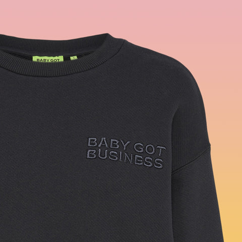 Schwarzer Sweater mit Rundhalsausschnitt und dezenter Ton-in-Ton-Stickerei "BABY GOT BUSINESS" auf der linken Brustseite.