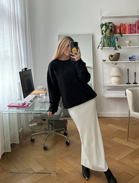Ann-Kathrin trägt den schwarzen "BABY GOT BUSINESS" Sweater kombiniert mit einem langen weißen Rock und schwarzen Boots in einem hell eingerichteten Homeoffice.